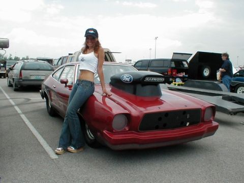 1978 Ford Mustang II - A woman poses with a modified red drag car a prominent hood scoop and custom grille.