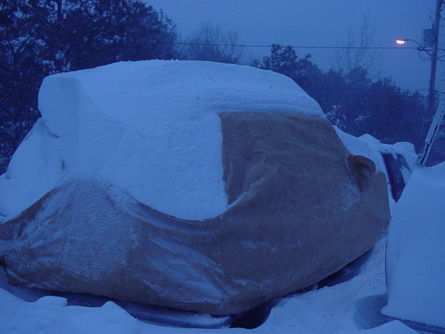 Car covered in snow with a tan tarp underneath, seen from a side angle