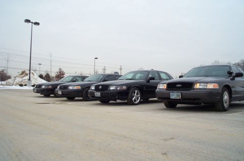 2001 Ford Crown Victoria - Four black Ford Crown Victorias are lined up in a parking lot on an overcast day.