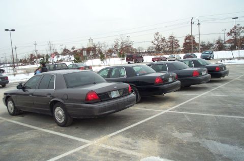 2001 Ford Crown Victoria - Several dark-colored Ford Crown Victorias are parked in a lot on an overcast day.
