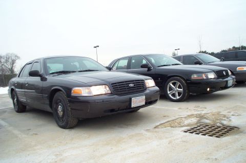 2001 Ford Crown Victoria - Two black Ford Crown Victorias are parked side-by-side in a lot, with a third vehicle partially visible behind them.