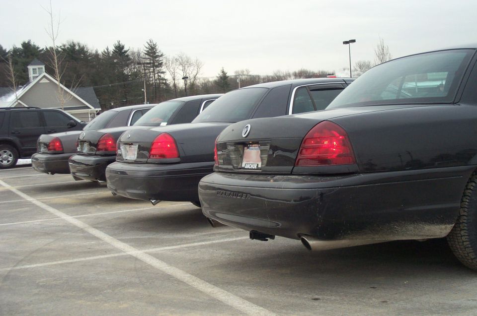 Four black Ford Crown Victorias parked in a lot, rear view. One SUV visible to the left