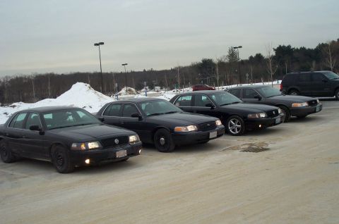 2001 Ford Crown Victoria - A line of four black 2001 Ford Crown Victoria sedans are parked in a lot with an SUV in the background.