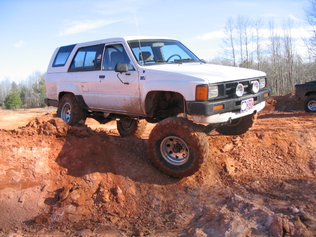 White Toyota 4Runner with off-road tires climbing a dirt hill. Another vehicle is visible in the background