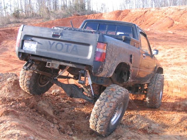 Dark gray Toyota pickup truck with large off-road tires climbing a dirt hill. Body style suggests 1980s