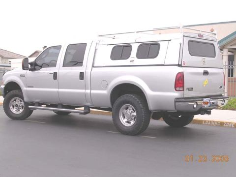 2004 Ford F-250 - A silver 2004 Ford F-250 pickup truck is parked on asphalt, equipped with a white camper shell and a roof rack.