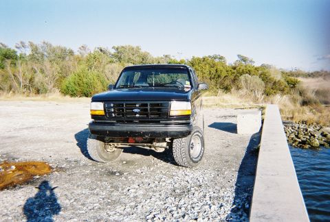 1996 Ford Bronco - Front view of a black 1996 Ford Bronco parked on a gravel lot next to a concrete structure and water.