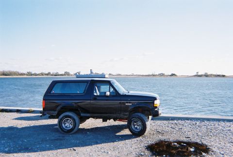 1996 Ford Bronco - A black Ford Bronco is parked on a gravel surface next to a body of water under a clear sky.