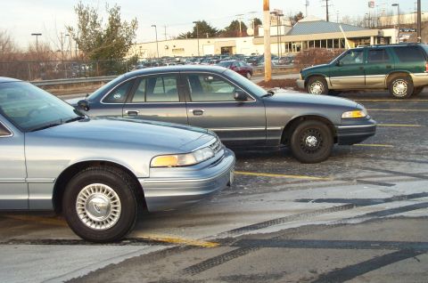 2001 Ford Crown Victoria - Two Ford Crown Victorias and a green SUV are parked in a lot with tire marks on the ground.