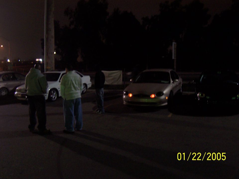 Silver sedan with orange turn signals parked at night. Two people stand nearby. Another car is visible to the right