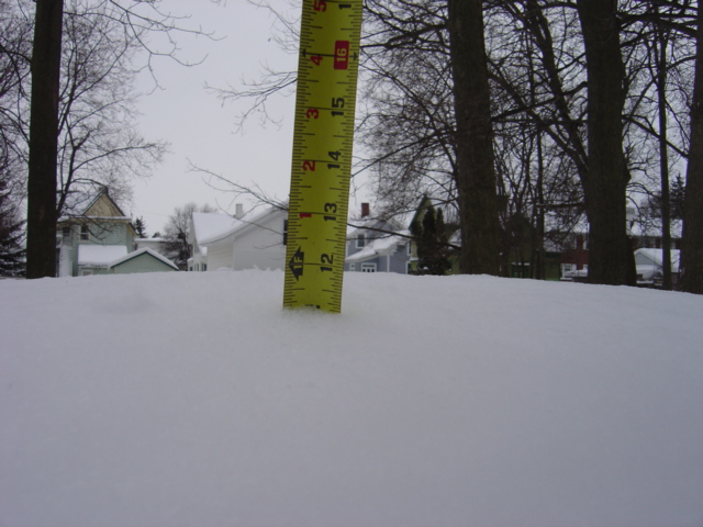 Yellow ruler inserted into snow, showing depth against a background of houses and trees