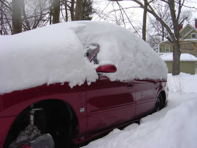 red car buried in snow, only side visible, trees and house in background
