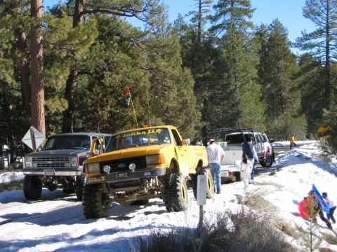 1985 Ford Bronco - Several modified trucks and SUVs are parked on a snowy road in a wooded area.