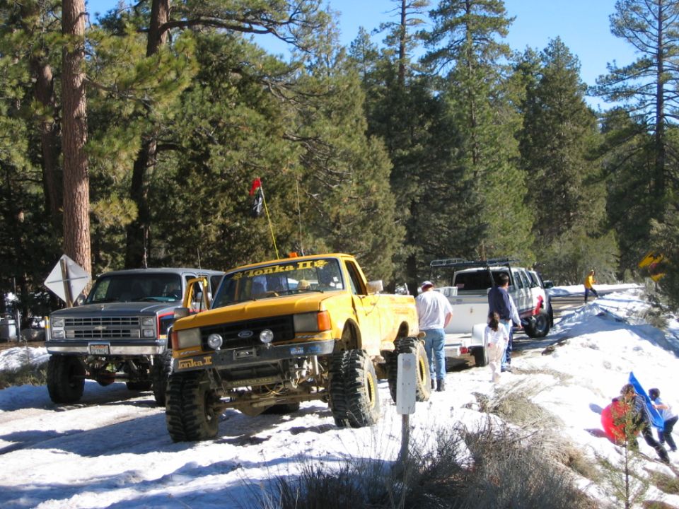 Yellow Ford pickup and black Chevy on big tires parked in snowy pine forest