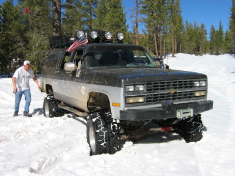 1985 Ford Bronco - Modified Chevrolet K5 Blazer with off-road tires stuck in the snow, a man is standing next to it.