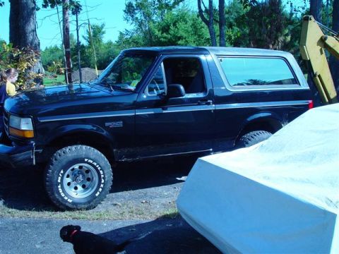 1996 Ford Bronco - A black Ford Bronco is parked next to a boat covered with a white tarp, with trees and a yellow excavator in the background.