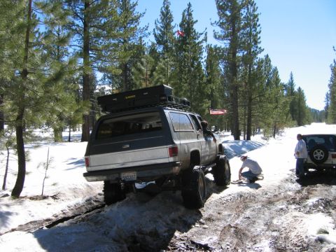 1985 Ford Bronco - Two SUVs navigate a snowy off-road path with trees surrounding them.