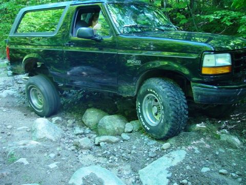 1996 Ford Bronco - A black Ford Bronco navigates a rocky path in a wooded area, with its front passenger side tire lifted.