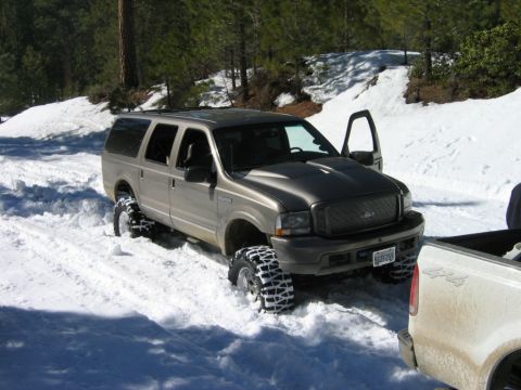 1985 Ford Bronco - A silver Ford Excursion is stuck in the snow with its driver door open, beside a white pickup truck.