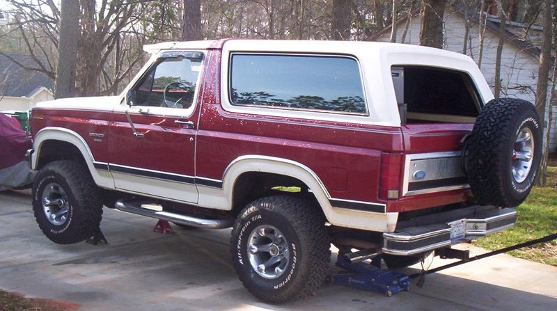 Red and white Ford Bronco SUV with lifted suspension and BFGoodrich tires, rear raised on a floor jack in a driveway