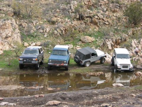 1989 Ford Bronco - Multiple off-road vehicles are parked near a muddy puddle in a rocky, outdoor environment.