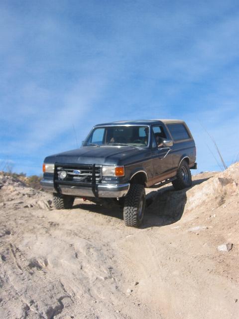 1989 Ford Bronco - A dark blue Ford Bronco navigates a rocky, off-road trail under a clear blue sky.