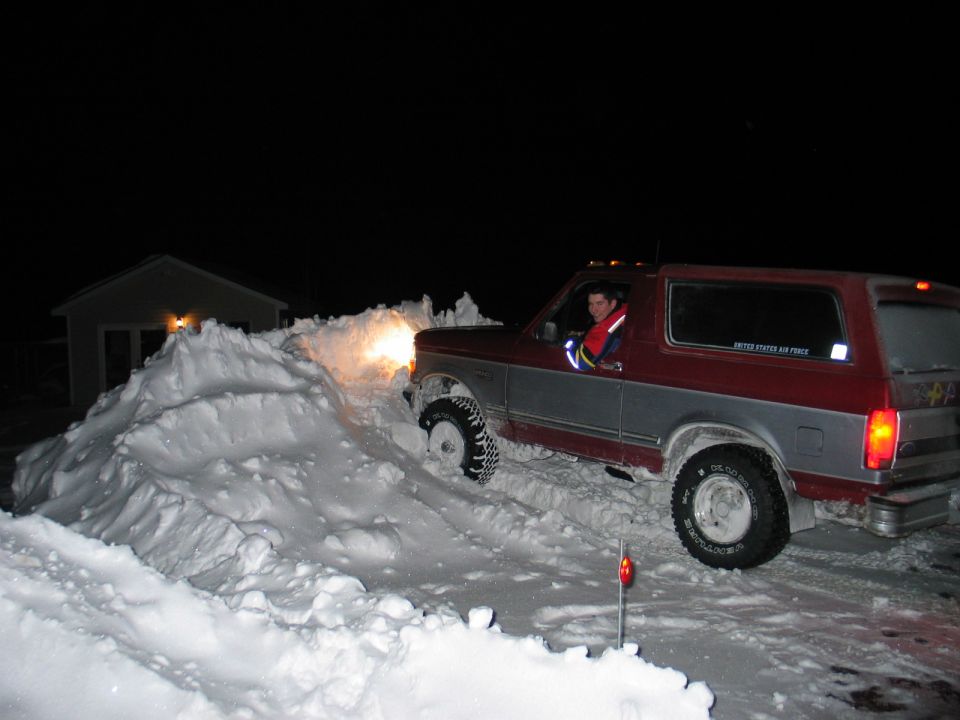 Maroon and silver Ford Bronco driving up a snow pile at night with driver visible