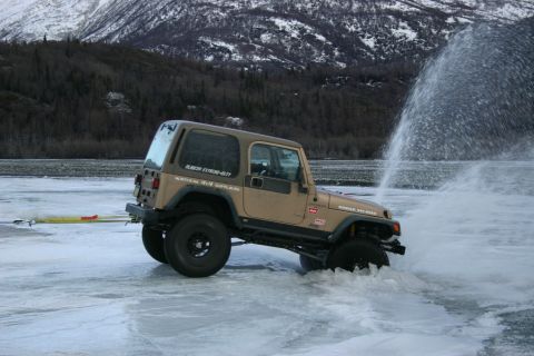 2003 Jeep TJ - A tan Jeep Wrangler TJ drives on ice, creating a large spray of water, with a snowy mountain range behind it.