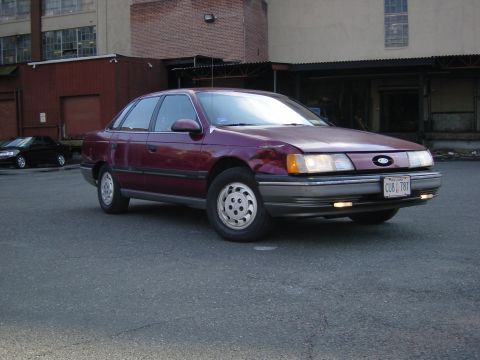 1989 Ford Taurus - Maroon 1989 Ford Taurus parked in front of a brick building. A black car is in the background.