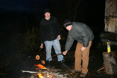 2003 Jeep TJ - Two men are cooking sausages over a campfire in a dark, wooded area.