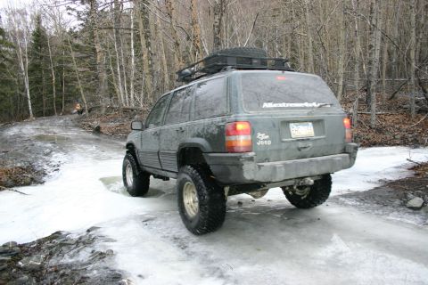 2003 Jeep TJ - A dark green Jeep Grand Cherokee navigates an icy path through a forest, equipped with a roof rack and spare tire.