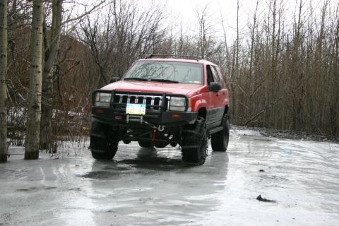 2003 Jeep TJ - A red Jeep Grand Cherokee equipped with an aftermarket bumper and oversized tires navigates an icy, wooded area.