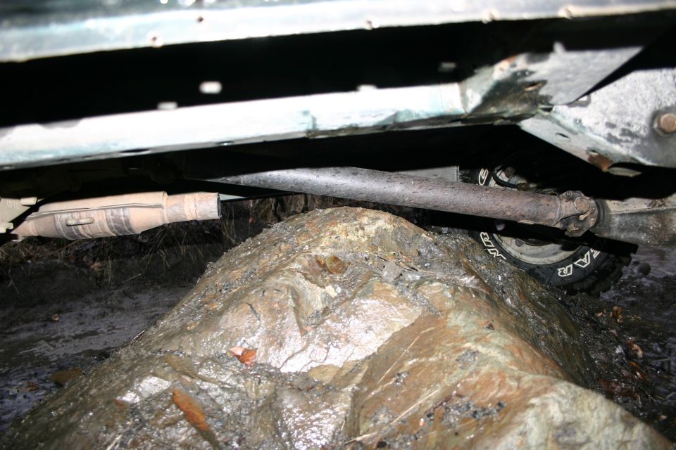 Vehicle undercarriage with driveshaft and tire visible, resting on a large rock