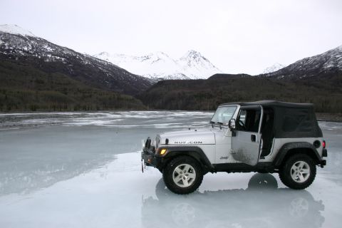 2003 Jeep TJ - A silver Jeep Wrangler is parked on a frozen body of water, with snow-capped mountains and dark trees behind it.