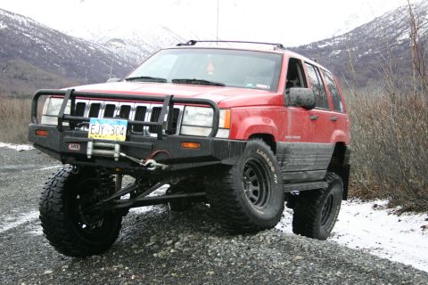 2003 Jeep TJ - This red Jeep Grand Cherokee is equipped with an ARB bumper and large off-road tires, parked on a gravel road with a snowy mountain landsca…