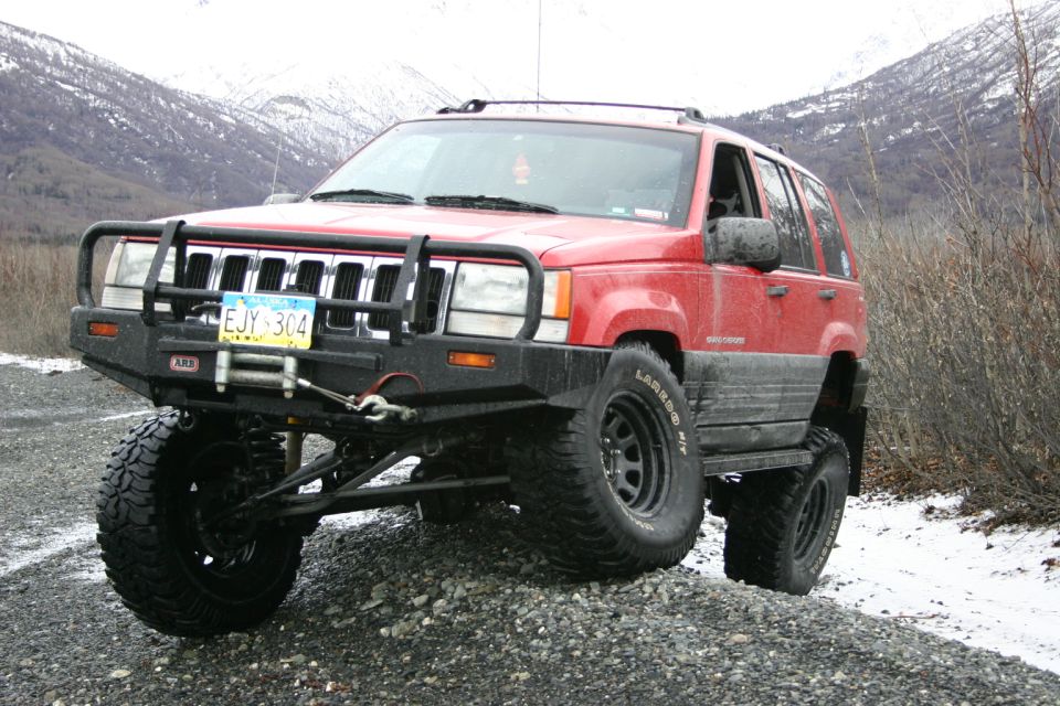 Red Jeep Grand Cherokee with aftermarket bumper and large tires on a gravel road with snowy mountains in background