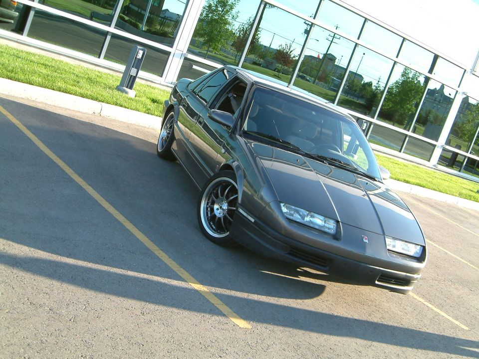 Dark gray Saturn sedan, front 3/4 view, black aftermarket wheels, parked by glass building