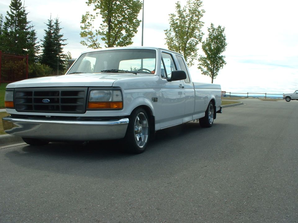 Lowered white Ford F-150 extended cab pickup with dark grille and polished wheels, front three-quarter view on a road