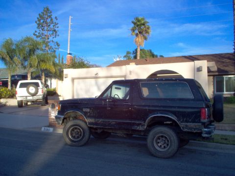 1988 Ford Bronco - A black Ford Bronco is parked on the street in front of a house, with another white Bronco visible in the driveway.