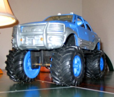 1978 Ford Bronco - A blue toy monster truck with large, knobby tires sits on a surface, with a lamp visible to the left.
