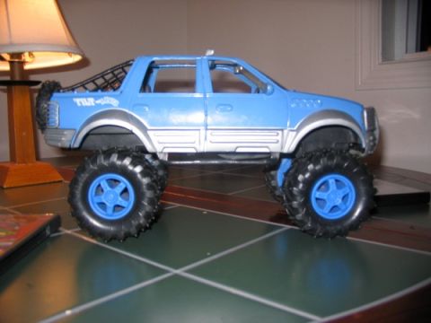 1978 Ford Bronco - A blue toy truck with oversized tires and a black roll bar is displayed on a surface with a grid pattern.