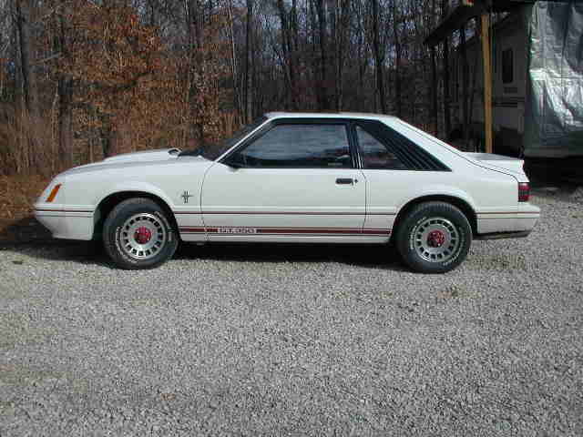 White Ford Mustang hatchback with red stripes, side view, parked on gravel near trees