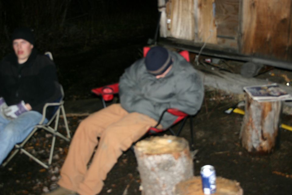 Two people sit in camping chairs outdoors at night next to a wooden structure. One person is asleep