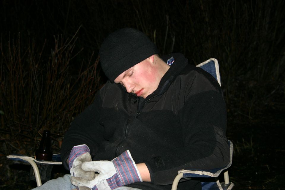 Person wearing a black beanie and jacket sleeps in a camping chair at night, with gloves on hands and a bottle nearby