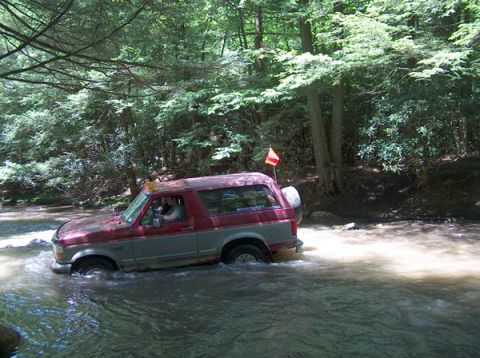 1995 Ford Bronco - A red and grey 1995 Ford Bronco navigates a stream in a forest, kicking up water as it goes.
