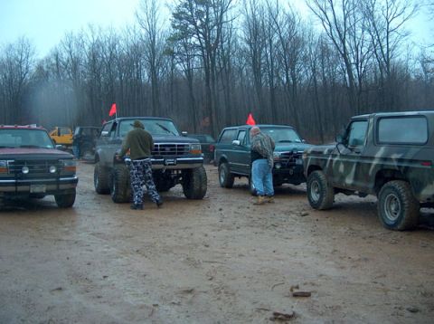 1995 Ford Bronco - Multiple off-road vehicles are gathered in a muddy, wooded area on an overcast day.
