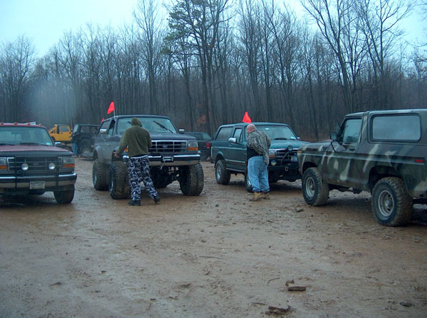 Several lifted trucks and SUVs parked on a muddy, wooded area. Two people are standing near the vehicles