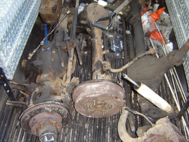 Several rusty vehicle axles and suspension components are piled in the bed of a truck