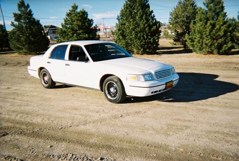 2000 Ford Crown Victoria - A white 2000 Ford Crown Victoria is parked on a dirt lot with trees and buildings in the background.