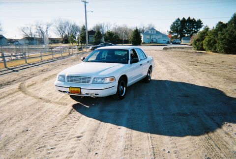 2000 Ford Crown Victoria - A white 2000 Ford Crown Victoria sedan is parked on a dirt road under a clear sky.